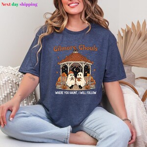 Puede incluir: Camiseta azul marino con el texto "Gilmore Ghouls" sobre una ilustraci&oacute;n de dos fantasmas en un mirador. La frase "WHERE YOU HAUNT, I WILL FOLLOW" est&aacute; impresa debajo. La camiseta tiene tem&aacute;tica de Halloween.