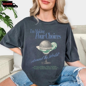 May include: Dark gray t-shirt with the text "I'm Making Pour Choices Around the World!" and a cocktail graphic. The graphic includes a cocktail glass with a golf ball and lime slice. The shirt also lists several countries.