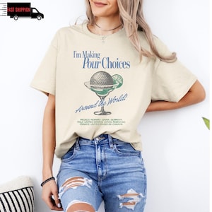 May include: Beige t-shirt with the text "I'm Making Pour Choices Around the World!" and a cocktail graphic. The graphic features a golf ball, lime, and a martini glass. The shirt also lists several countries.