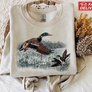 Pu&ograve; includere: Una felpa beige con una grafica dettagliata di un germano reale in volo, con altre due anatre sullo sfondo. L'opera d'arte comprende sfumature di marrone, verde e blu, su un paesaggio di acqua e canneti. La felpa ha un girocollo.