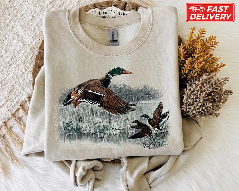 Pu&ograve; includere: Una felpa beige con una grafica dettagliata di un germano reale in volo, con altre due anatre sullo sfondo. L'opera d'arte comprende sfumature di marrone, verde e blu, su un paesaggio di acqua e canneti. La felpa ha un girocollo.