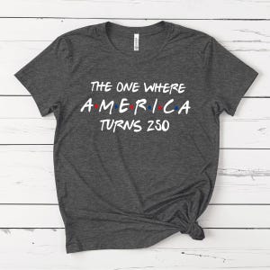 May include: A dark heather gray short-sleeve t-shirt with white text that reads THE ONE WHERE AMERICA TURNS 250. The word AMERICA features small red and blue dots between the letters, mimicking a classic television show logo style.
