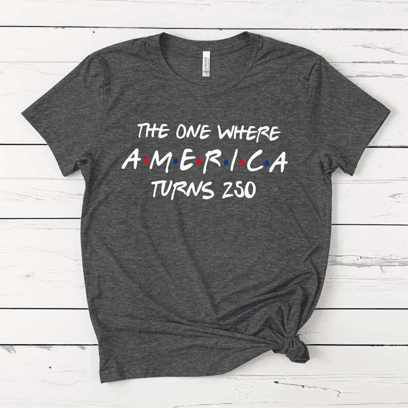 May include: A dark heather gray short-sleeve t-shirt with white text that reads THE ONE WHERE AMERICA TURNS 250. The word AMERICA features small red and blue dots between the letters, mimicking a classic television show logo style.