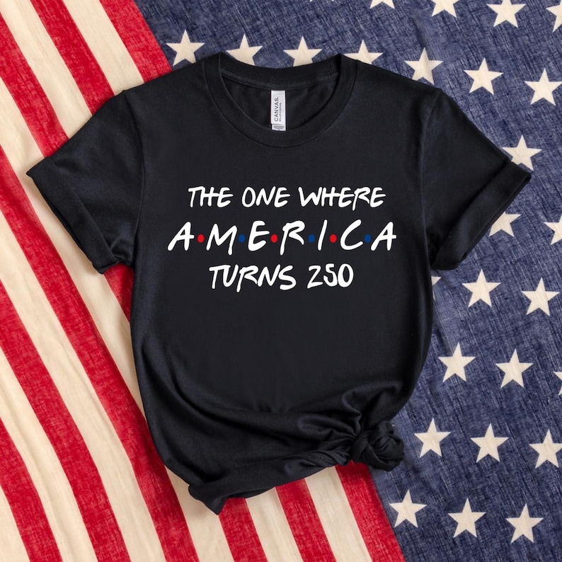 May include: A black short-sleeved t-shirt with white text that reads THE ONE WHERE AMERICA TURNS 250. The word AMERICA features small red and blue dots between the letters. The shirt has a crew neckline and a standard fit with a visible brand label.