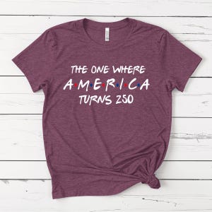 May include: A heathered maroon short-sleeve t-shirt with white screen-printed text that reads THE ONE WHERE AMERICA TURNS 250, featuring small red and blue dots between the letters of the word AMERICA, and a white brand label at the inner neck.