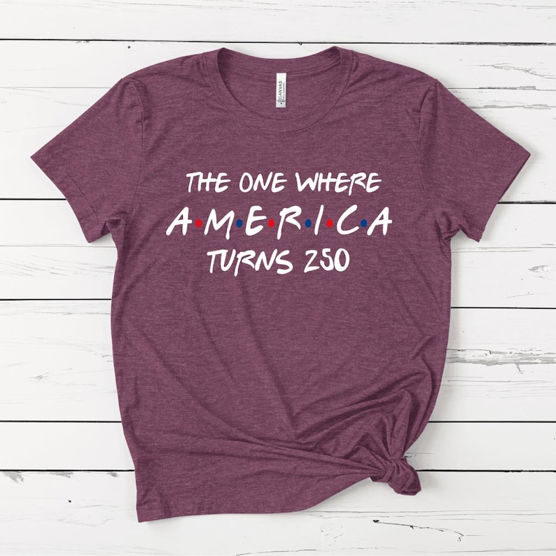 May include: A heathered maroon short-sleeve t-shirt with white screen-printed text that reads THE ONE WHERE AMERICA TURNS 250, featuring small red and blue dots between the letters of the word AMERICA, and a white brand label at the inner neck.