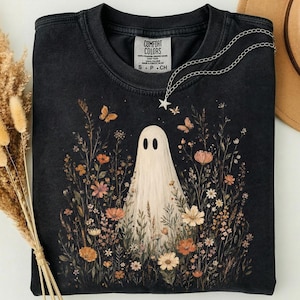 May include: A black short-sleeve t-shirt featuring a graphic of a white ghost surrounded by various orange, pink, and white wildflowers and small butterflies. The shirt has a crew neckline and a Comfort Colors brand label printed on the inside neck.