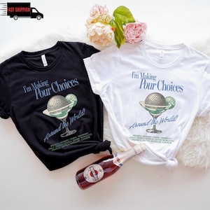 May include: Two t-shirts, one black and one white, with the text "I'm Making Pour Choices Around the World!" and a cocktail graphic. A bottle of red sparkling wine is in the foreground. Floral arrangement in the background.