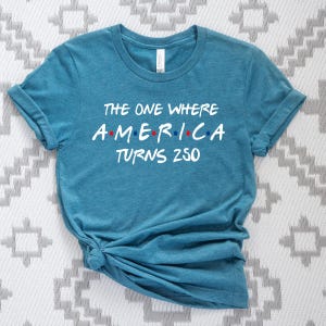 May include: A heathered teal short-sleeve t-shirt featuring white text that reads THE ONE WHERE AMERICA TURNS 250. The word AMERICA is stylized with red and blue dots between the letters, mimicking a classic television show logo design.