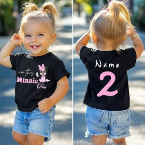 以下が含まれることがあります： ミニーマウスのグラフィックとピンク色の「I'm this Minnie」という文字が入った黒いTシャツ。Tシャツの背面には「NAME」という文字と数字の「2」がピンク色でプリントされています。子供は青いデニムのショートパンツを着用しています。