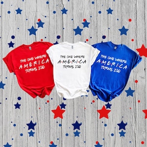 May include: Three short-sleeved t-shirts in red, white, and blue, each featuring the black printed text THE ONE WHERE AMERICA TURNS 250, with the word AMERICA stylized with dots between each letter, arranged horizontally on a wooden surface.