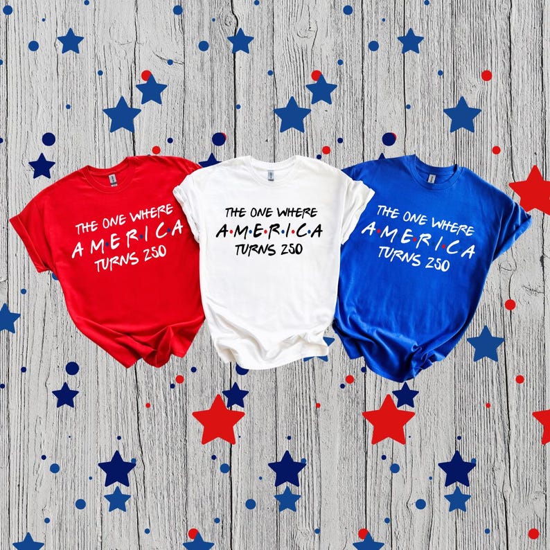 May include: Three short-sleeved t-shirts in red, white, and blue, each featuring the black printed text THE ONE WHERE AMERICA TURNS 250, with the word AMERICA stylized with dots between each letter, arranged horizontally on a wooden surface.