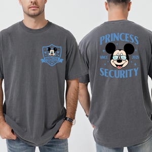 May include: Gray t-shirt featuring a Mickey Mouse graphic with the text "PRINCESS SECURITY" in blue. The front of the shirt has a shield design with Mickey Mouse. The back of the shirt has a larger Mickey Mouse graphic with the text "PRINCESS SECURITY SINCE 2025".