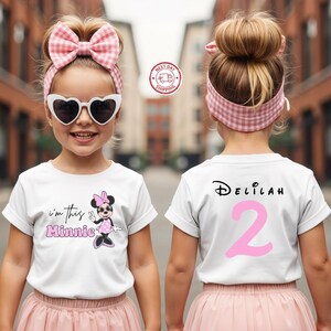 以下が含まれることがあります： ピンクのアクセントが付いた2つの白いTシャツ。前面のシャツにはミニーマウスのグラフィックと「I'm this Minnie」というテキストがあります。背面のシャツには「Delilah」という名前と数字の「2」があります。どちらのシャツも子供用です。