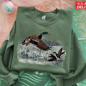 Pu&ograve; includere: Una felpa verde oliva con una grafica dettagliata di un germano reale in volo, con altre due anatre sullo sfondo. L'opera d'arte &egrave; ambientata su uno sfondo di canne e acqua, creando un design a tema natura.