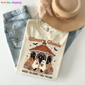 Puede incluir: Camiseta color crema con el texto "Gilmore Ghouls" y un gr&aacute;fico de Halloween con fantasmas, calabazas y un mirador. La camiseta se combina con vaqueros azules y un sombrero color canela.