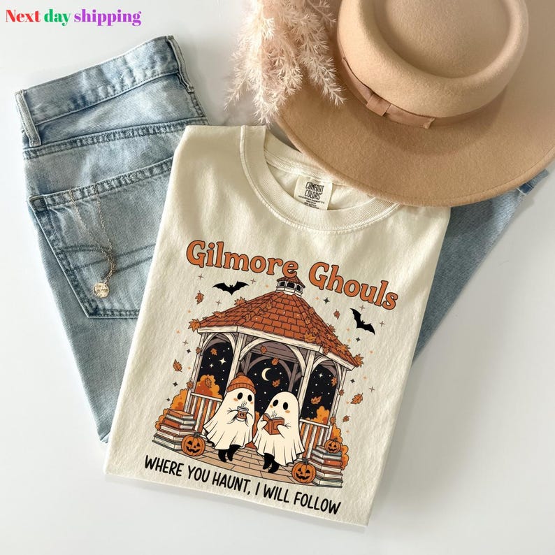 Puede incluir: Camiseta color crema con el texto "Gilmore Ghouls" y un gr&aacute;fico de Halloween con fantasmas, calabazas y un mirador. La camiseta se combina con vaqueros azules y un sombrero color canela.