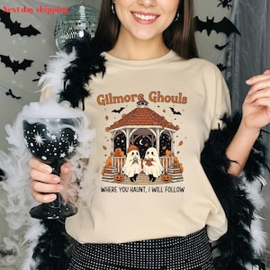 Puede incluir: Camiseta beige con un dise&ntilde;o de Halloween. El dise&ntilde;o presenta una glorieta con dos figuras de fantasmas, calabazas y el texto "Gilmore Ghouls, Where You Haunt, I Will Follow". Murci&eacute;lagos tambi&eacute;n est&aacute;n incluidos en el dise&ntilde;o.