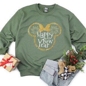 Pu&ograve; includere: Una felpa verde con un design glitterato dorato delle orecchie di Topolino e la scritta "Happy New Year". L'immagine include anche una piccola confezione regalo con un nastro rosso e una composizione floreale festosa.