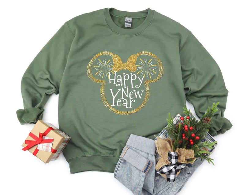 Pu&ograve; includere: Una felpa verde con un design glitterato dorato delle orecchie di Topolino e la scritta "Happy New Year". L'immagine include anche una piccola confezione regalo con un nastro rosso e una composizione floreale festosa.