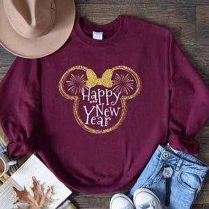 Pu&ograve; includere: Una felpa bordeaux con un design glitterato dorato a forma di testa di Topolino con un fiocco e le parole "Happy New Year" in bianco. La felpa &egrave; abbinata a jeans blu e scarpe da ginnastica bianche.