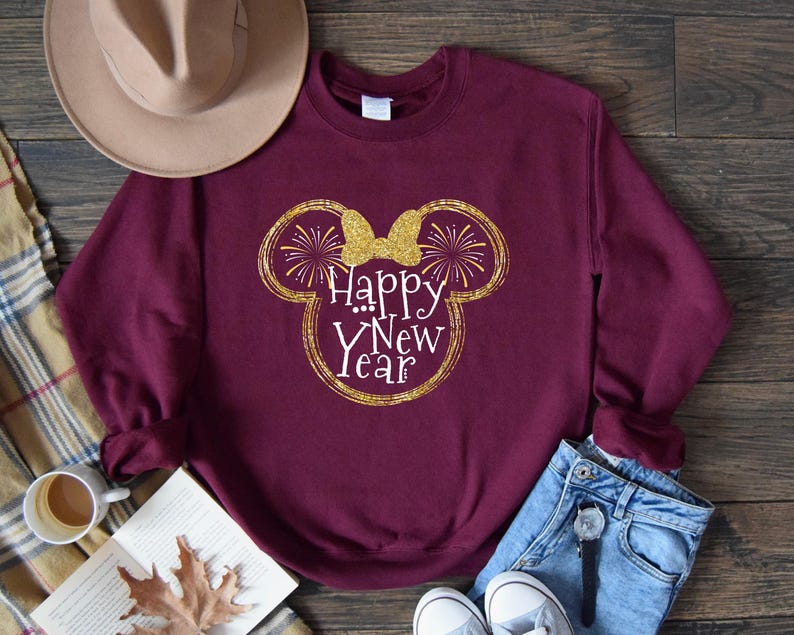 Pu&ograve; includere: Una felpa bordeaux con un design glitterato dorato a forma di testa di Topolino con un fiocco e le parole "Happy New Year" in bianco. La felpa &egrave; abbinata a jeans blu e scarpe da ginnastica bianche.