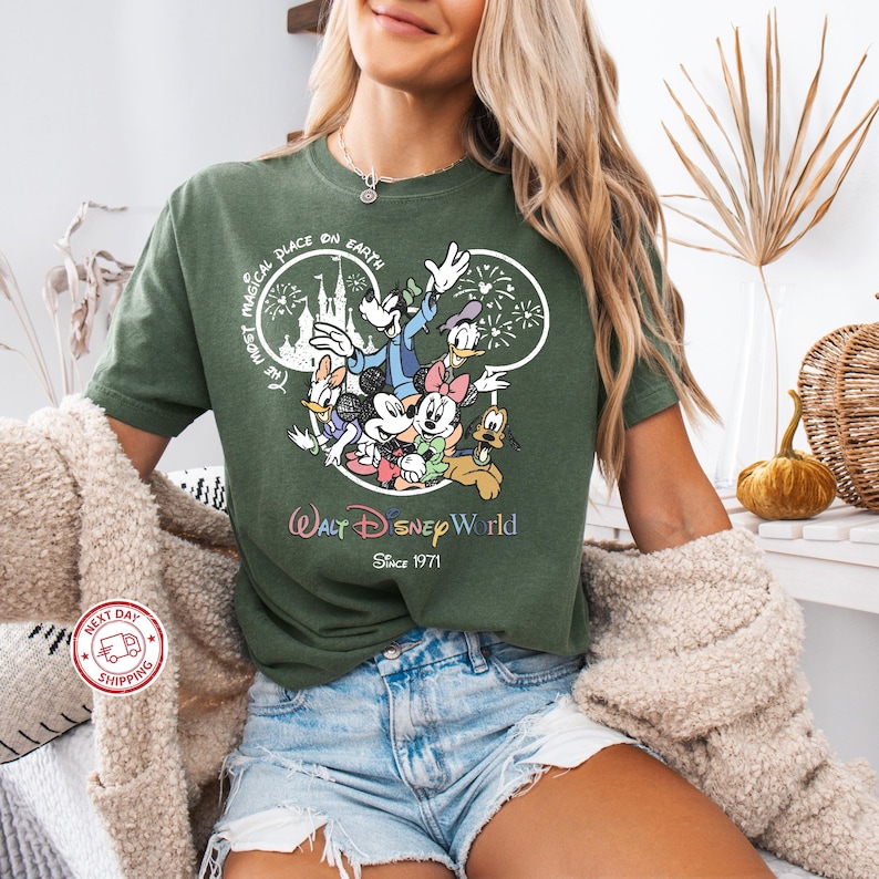 Puede incluir: Camiseta verde oscuro con un gr&aacute;fico colorido de personajes de Disney, incluyendo Mickey y Minnie Mouse, Donald Duck y Goofy. La camiseta tiene el texto "Walt Disney World" y "Since 1971". La frase "The most magical place on Earth" tambi&eacute;n est&aacute; impresa.