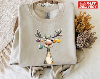 Puede incluir: Sudadera beige con una ilustración caprichosa de un reno. El reno tiene grandes cuernos adornados con adornos coloridos. El diseño es de estilo caricaturesco, perfecto para la temporada navideña. La sudadera está doblada sobre una superficie texturizada.