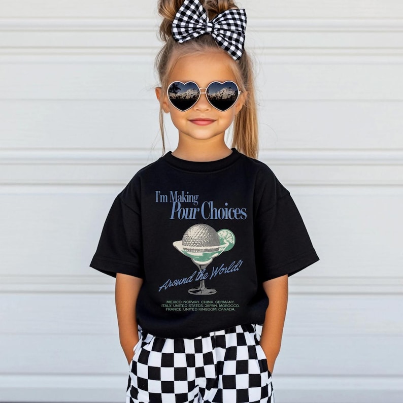 May include: A black t-shirt with the text "I'm Making Pour Choices Around the World!" and a graphic of a cocktail. The shirt is worn by a child wearing heart-shaped sunglasses and a black and white checkered bow.