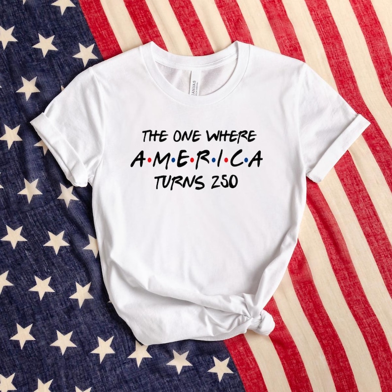 May include: A white short-sleeved crew neck t-shirt featuring black text that reads THE ONE WHERE AMERICA TURNS 250, with the word AMERICA stylized with colorful dots between the letters in a font reminiscent of a popular television show logo.