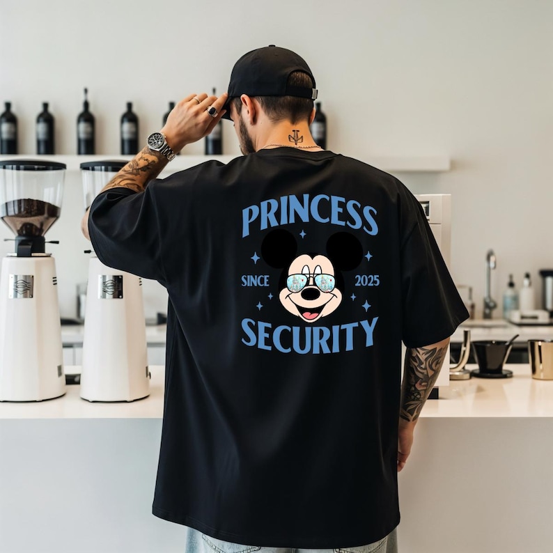 May include: Black t-shirt with a graphic of Mickey Mouse wearing glasses and the words "PRINCESS SECURITY SINCE 2025" in blue. The shirt is a casual, oversized fit.