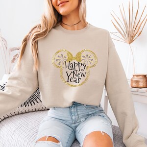 Pu&ograve; includere: Felpa beige con un design a testa di Topolino glitterato dorato. Il design include le parole "Happy New Year" in corsivo nero. La felpa ha maniche lunghe e girocollo.