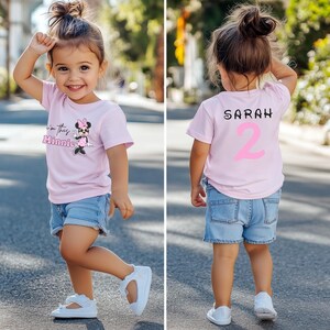 以下が含まれることがあります： 薄いピンク色のTシャツとデニムのショートパンツを着た幼い子供。Tシャツの前面にはミニーマウスのグラフィックと「I'm this Minnie」の文字があります。Tシャツの背面には「SARAH」という名前とピンク色の数字「2」があります。