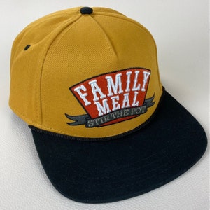 May include: A mustard yellow baseball cap with a black brim. The front of the cap features an embroidered patch with the text "Family Meal Stir the Pot" in white and red lettering.