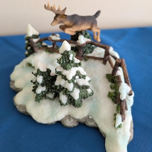May include: A decorative figurine featuring a brown moose leaping over a brown wooden fence. The scene includes green pine trees covered in white snow, set upon a textured, snow-covered base that mimics a winter landscape with icy blue accents.