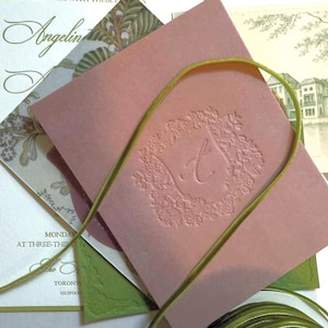 May include: Wedding stationery set with a dusty pink envelope embossed with floral patterns and the letter "A". Other cards feature floral and architectural designs, tied with a green ribbon. Text includes "Together with their families" and "Angelina".