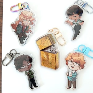May include: A selection of acrylic keyrings featuring cartoon characters and a gold lighter. The keyrings depict various designs, including a character with long curly hair, a character in a Hawaiian shirt, and a character with glasses.