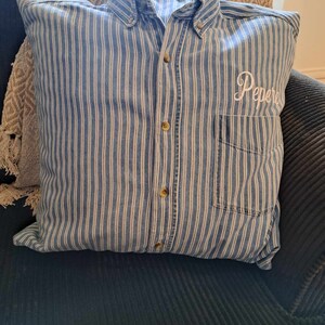May include: A blue and white striped pillow with a button-down collar and a pocket. The pillow has the word "Pepere" embroidered on it.