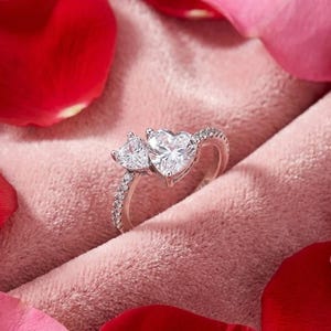 May include: A silver ring featuring two heart-shaped gemstones, one larger than the other, set on a pink velvet surface. The band is encrusted with small, clear stones. Red and pink rose petals surround the ring.