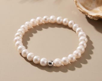 May include: A circular bracelet composed of numerous small, off-white, round pearls strung together in a continuous loop. A single, polished, reflective silver-toned metal bead is positioned prominently among the pearls, creating a focal point.