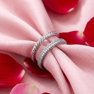 May include: Two silver rings sit on a pink cloth, with red rose petals scattered around. One ring features a twisted rope pattern, the other is set with small, sparkling stones. These rings are likely for a wedding or engagement.