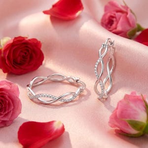 May include: Silver hoop earrings with an interwoven design, set with small, sparkling stones. The earrings are presented on a pink satin surface, with red rose petals and pink roses, creating a romantic scene.