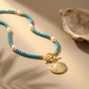 May include: A necklace featuring small turquoise beads interspersed with white pearls and gold-toned metal beads. The piece includes a gold-toned toggle clasp and a gold-toned seashell pendant embellished with small, clear, sparkling stones.