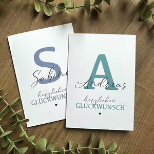 Può includere: Due biglietti bianchi con testo e nomi in tedesco. Un biglietto presenta una grande lettera "S" blu e il nome "Sabine". L'altro biglietto ha una "A" turchese e il nome "Andreas". Entrambi i biglietti hanno la frase "herzlichen GLÜCKWUNSCH".