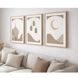 May include: Three framed art prints with a beige and white color scheme. The prints feature minimalist illustrations of a mosque, hanging lanterns, and a crescent moon over rolling hills. The frames are a light wood color.