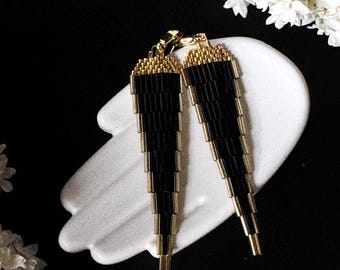 Pode incluir: Um par de brincos de contas pretas com detalhes dourados. Os brincos são exibidos em um prato branco em forma de mão contra um fundo preto, com flores brancas nos cantos.