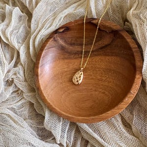 Puede incluir: Un collar de oro con un delicado colgante calado en forma de l&aacute;grima. El collar se muestra en un cuenco de madera y un fondo de tela beige.