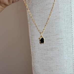 May include: A gold-colored necklace with a small, square, black pendant. The pendant is set in a gold frame and hangs from a delicate chain. The necklace is displayed on a neutral-colored background.