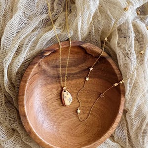 Puede incluir: Dos collares de oro en un cuenco de madera. Uno de los collares tiene un peque&ntilde;o colgante ovalado con un dise&ntilde;o de estrella. El otro collar tiene peque&ntilde;os abalorios en forma de estrella.