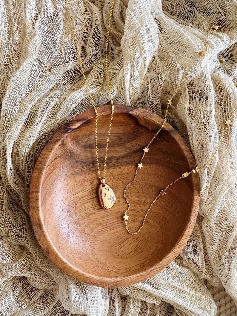 Puede incluir: Dos collares de oro en un cuenco de madera. Uno de los collares tiene un peque&ntilde;o colgante ovalado con un dise&ntilde;o de estrella. El otro collar tiene peque&ntilde;os abalorios en forma de estrella.